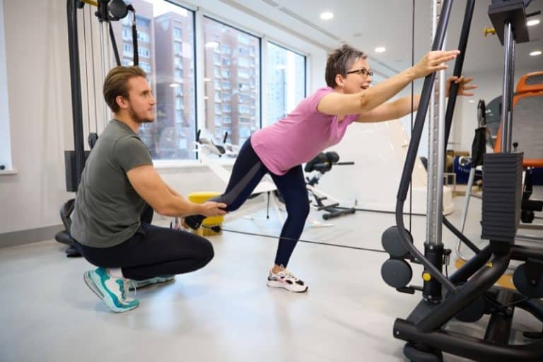 Male trainer helping adult woman doing sport exercise for leg in gym