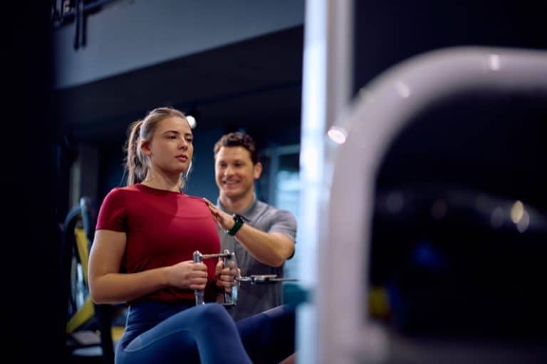 Personal trainer assisting athletic woman in exercising on seated cable row machine in a gym